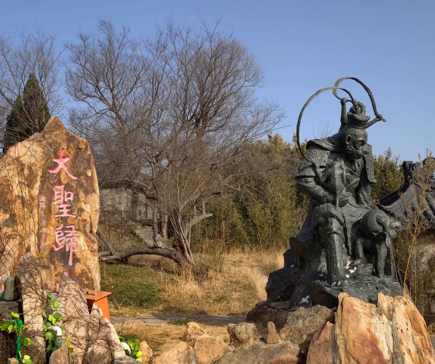 Bronze statue of a mythic Chinese warrior holding a circular weapon, perched on rocks beside a rock with red Chinese inscriptions in a park setting with leafless trees nearby.