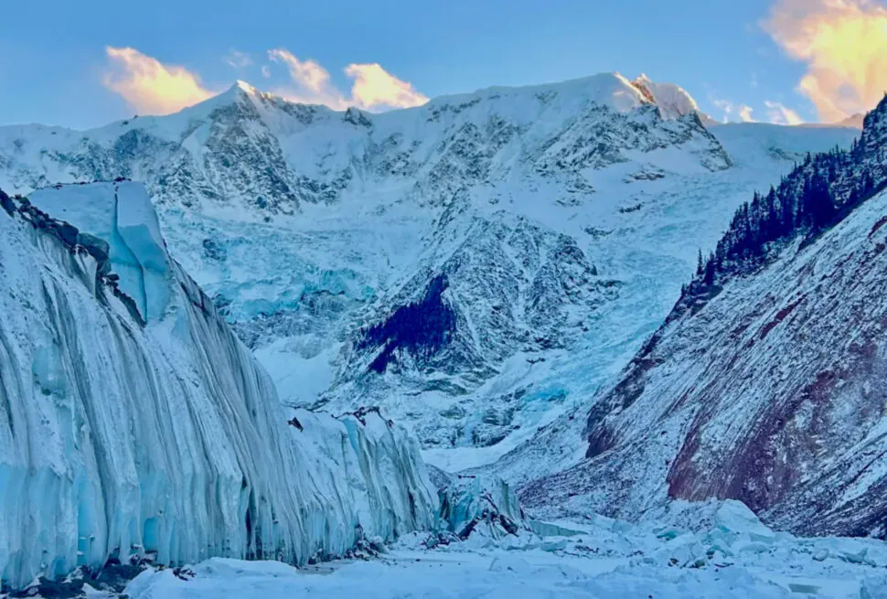 Snowy glacier with tall ice seracs and jagged mountain peaks under a blue sky at sunset.