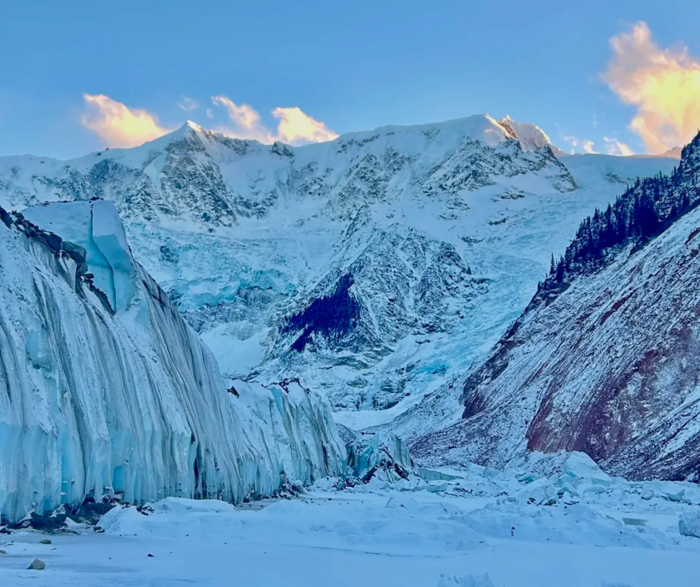 Snowy glacier with tall ice seracs and jagged mountain peaks under a blue sky at sunset.