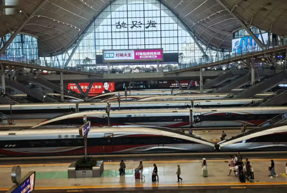 High-speed trains at a modern train station in China with a large glass roof and digital signage.