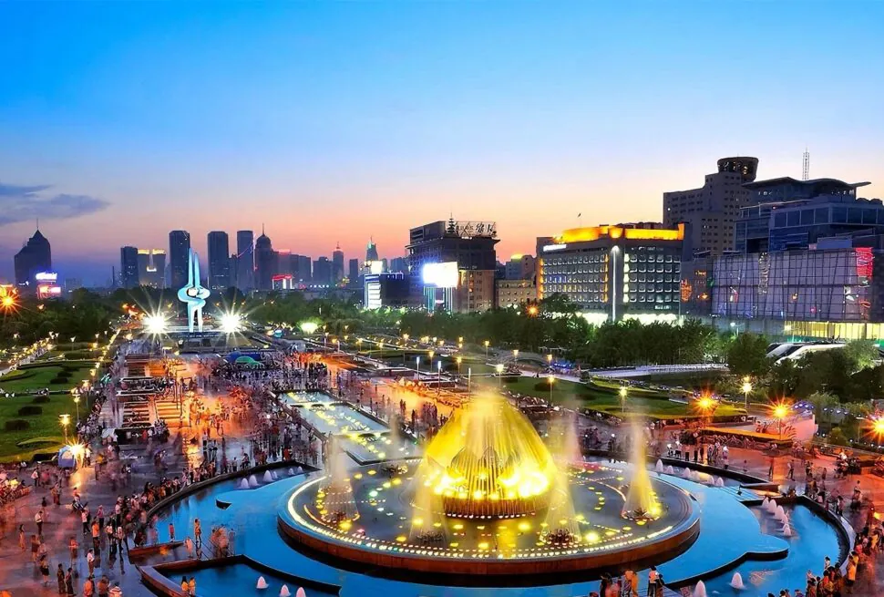 Night view of a city square with a large illuminated fountain and modern buildings.