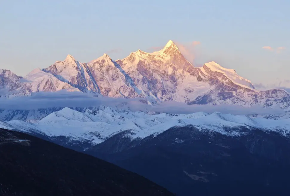 Snowy mountain range bathed in pink sunrise light, with dark forested valleys in the foreground.