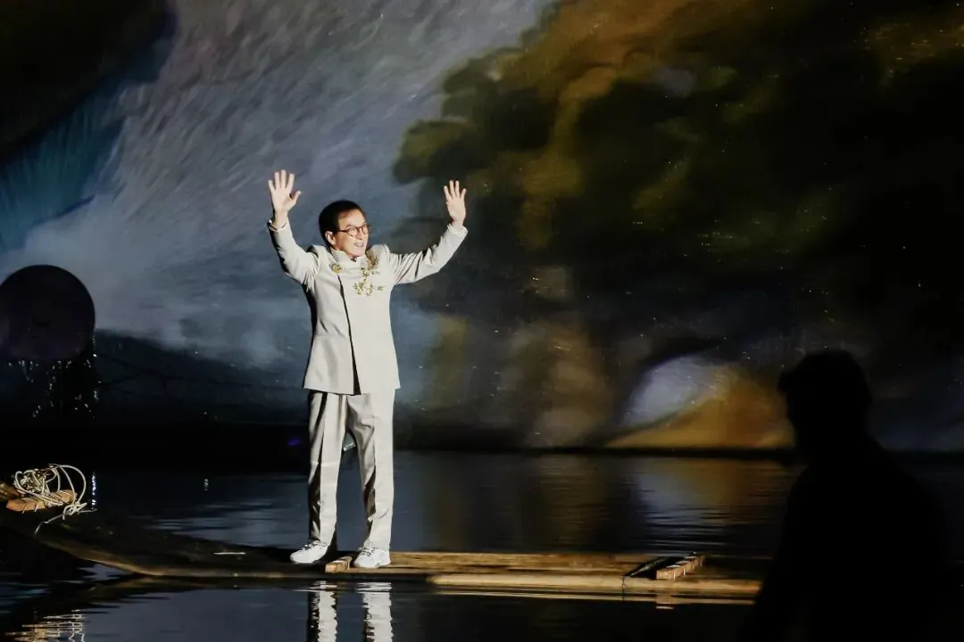 Man in a light gray suit stands on a wooden raft at night, arms raised as a colorful projection fills the backdrop behind him.