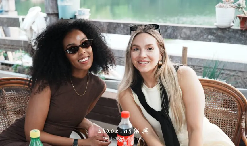 Two women smiling at a waterfront outdoor cafe with bottled drinks on the table.