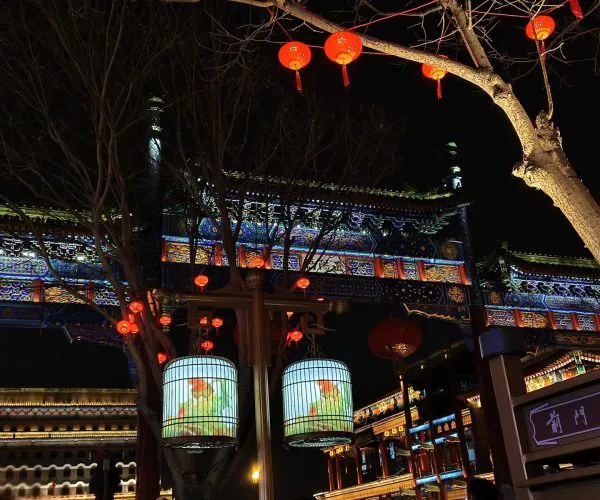 beijing, chinatravel, Night view of a traditional Chinese temple gate with red lanterns, colorful illuminated eaves, and bare trees in the foreground.