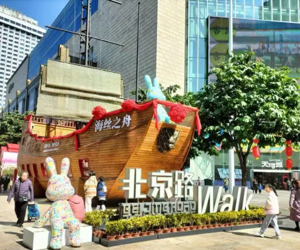Large wooden ship sculpture decorated with red ribbons sits in a busy city square, with a colorful bunny statue and a big foreground sign reading “Walk” near shoppers and modern buildings.