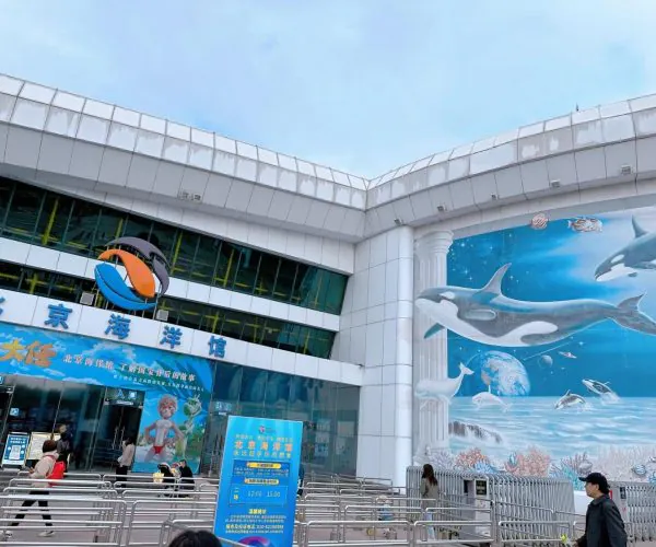Beijing Aquarium, Beijing, chinatravel, Entrance to Beijing Aquarium with a large blue mural of dolphins and sea life on the right, Chinese signage overhead, and people walking outside.