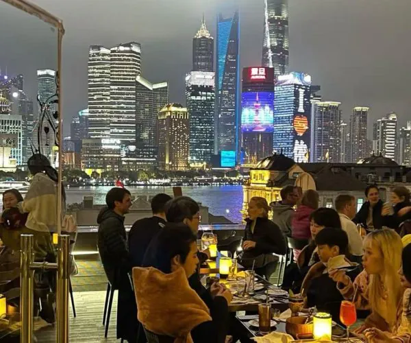 Diners seated at an outdoor rooftop restaurant at night, with a glowing city skyline and river in the background.