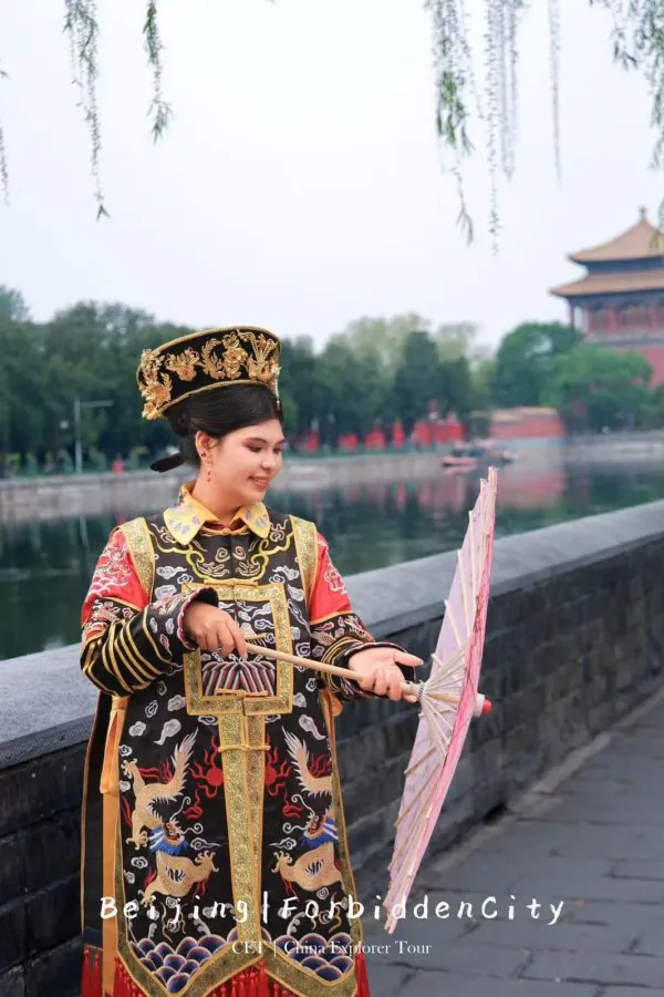 hanfu photoshoot, the forbidden city, beijing, qing dynasty, female, solo, chinatrip, Woman in an elaborate traditional Chinese imperial costume and headdress, holding a pink parasol beside a stone railing by a waterway, with a pavilion-style building in the background.