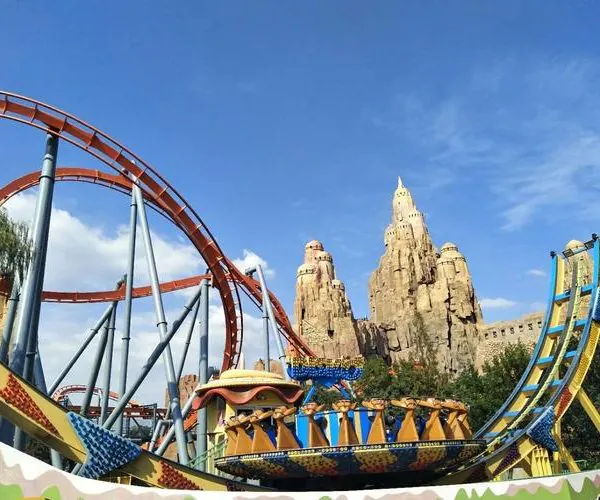 Beijing Happy Valley, Beijing, chinatravel, Colorful roller coaster with orange tracks looping around blue supports beside a rocky, castle-like structure on a sunny day at a theme park.