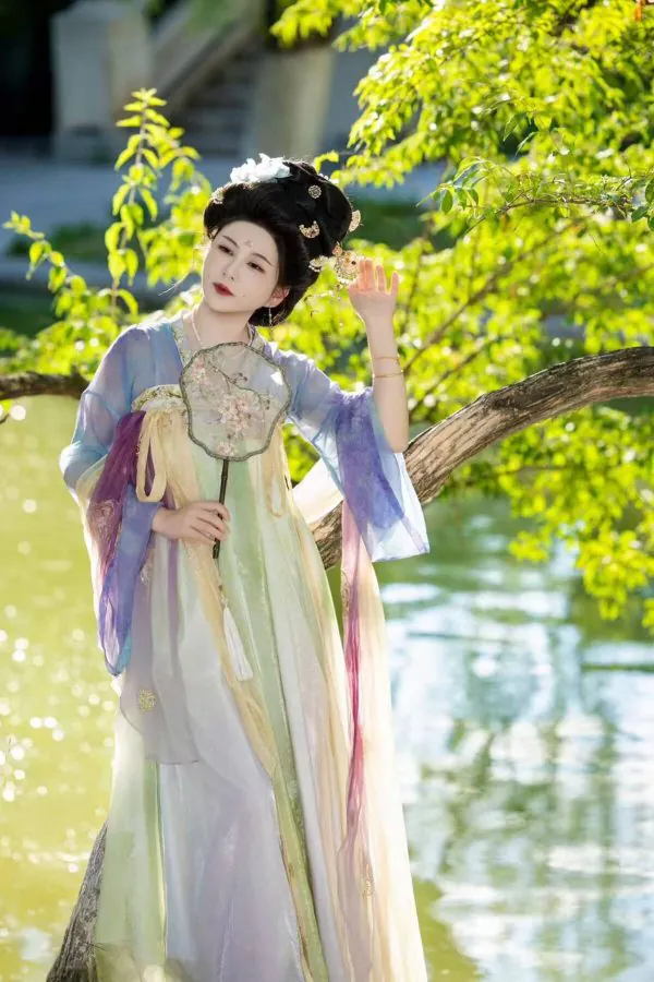 Hanfu photoshoot, zhongshan park, beijing, solo, tang dynasty, female, chinatravel, Woman in a pastel hanfu with an elaborate updo stands beside a tree by a sunlit pond, holding a delicate round floral fan and looking to the side.