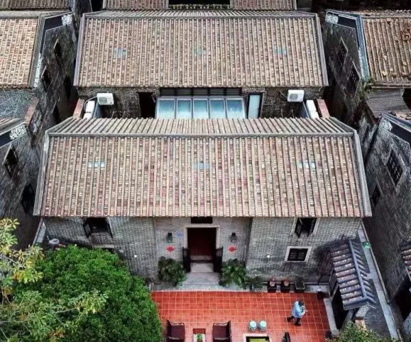 Aerial view of grey brick houses with tiled roofs; a red-tiled courtyard sits in front of a central house, with a person standing near the doorway.