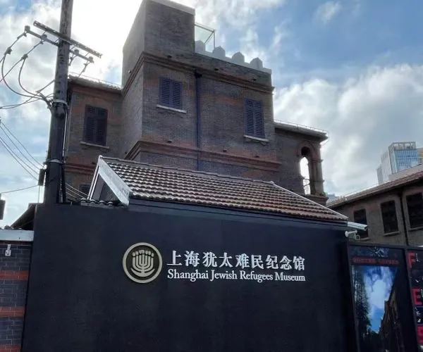 Exterior view of the Shanghai Jewish Refugees Museum with a dark wall sign displaying the museum name in Chinese and English behind a brick building.