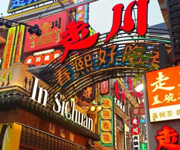 Neon-lit Chinatown street with colorful signs and an arched entrance, filled with Chinese characters and storefronts.