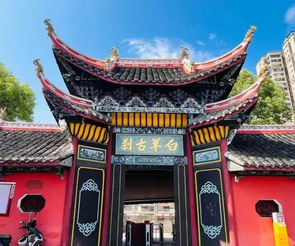 Ornate Chinese temple gate with red walls, black and gold accents, and curved tiled roofs, set against a blue sky and city buildings in the background.