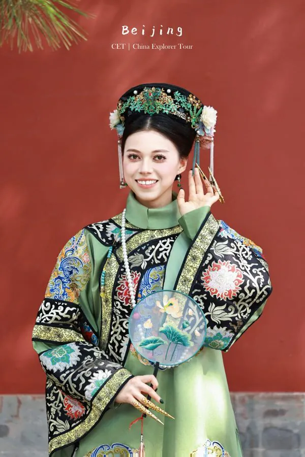 hanfu photoshoot, the forbidden city, beijing, qing dynasty, female, solo, chinatravel, Smiling woman in an ornate green and black traditional Chinese costume with a jeweled headdress, holding a round painted fan against a red backdrop.