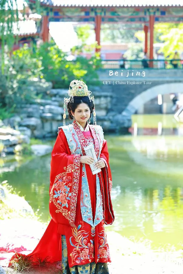 Woman in an ornate red Chinese imperial dress and gold headdress stands by a pond with a stone bridge in a lush garden, Beijing tourism imagery