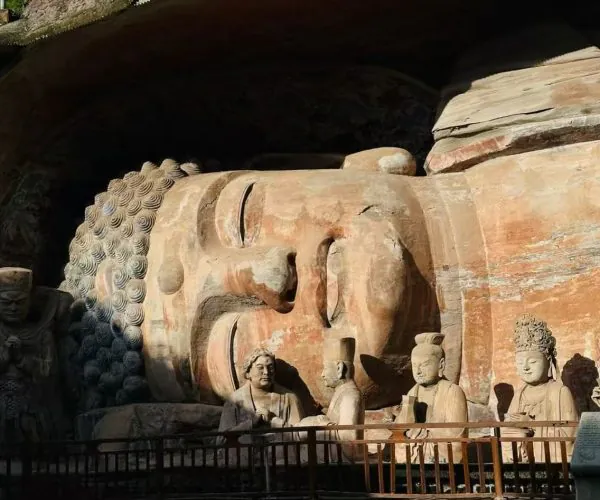 Large reclining Buddha carved in rock inside a cave, with several standing and seated stone figures nearby and a railing in front.