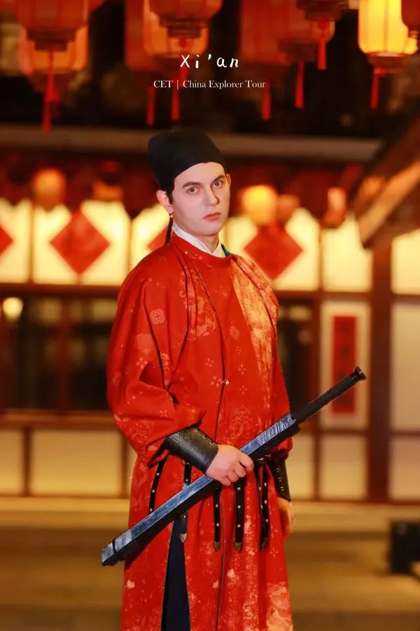 Hanfu photoshoot, xi'an, xingqing palace, male, chinatravel, Person in red traditional Chinese robe with a sword, standing in a lantern-lit setting; overlay text reads 'Xi’an CET | China Explorer Tour'