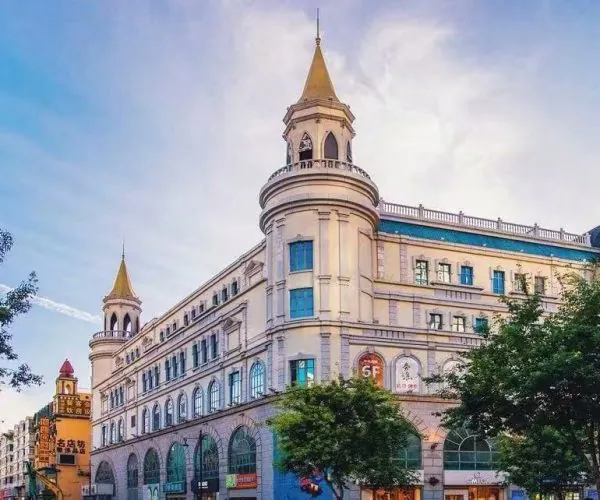 Historic European-style shopping arcade with two tower spires and arched storefronts along a tree-lined street on a sunny day.