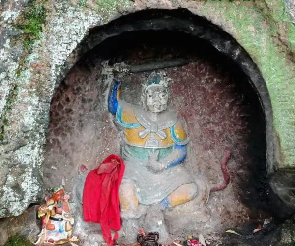 Statue of a seated deity carved into a rocky cave, with yellow and blue clothing, a red cloth draped nearby, and small figurines around the altar in a moss-covered alcove.