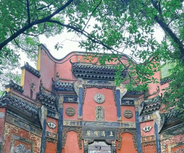 Ornate red Chinese temple gate with blue-gray decorative panels and gold carvings, framed by green trees.