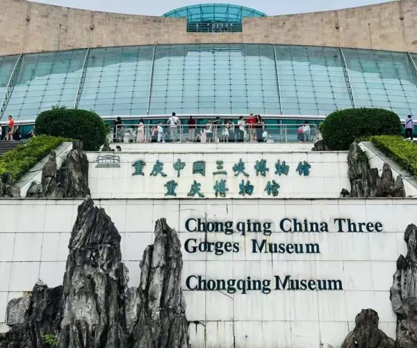 Exterior view of Chongqing China Three Gorges Museum with a glass-domed atrium, visitors on the terrace, and rock sculptures framing a sign that reads 'Chongqing China Three Gorges Museum' in English and Chinese.