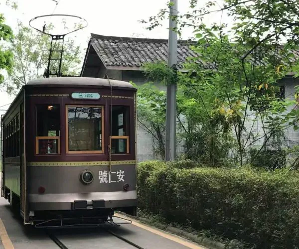 Vintage maroon tram car on a narrow rail line, flanked by trees and a hedge with a brick building in the background.