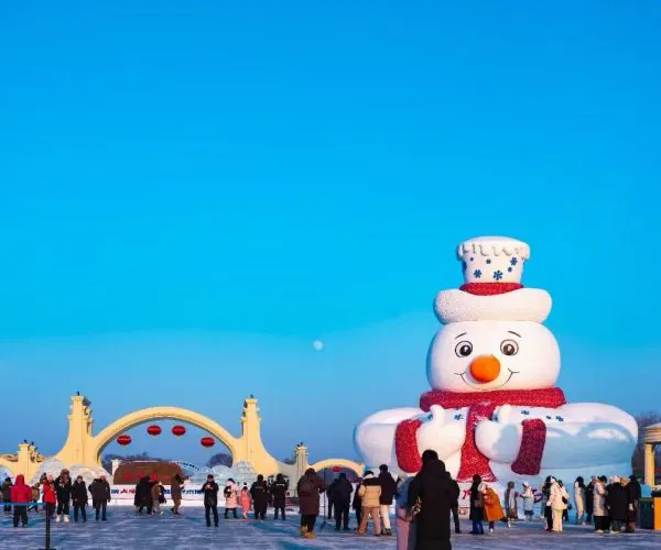 Large inflatable snowman sculpture with red scarf and hat in a frozen plaza, crowds skating below under a clear blue sky.