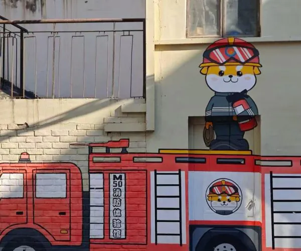 Colorful mural of a red fire truck painted on a wall with a cartoon firefighter cat standing on top beside a window and ladders drawn on the side.