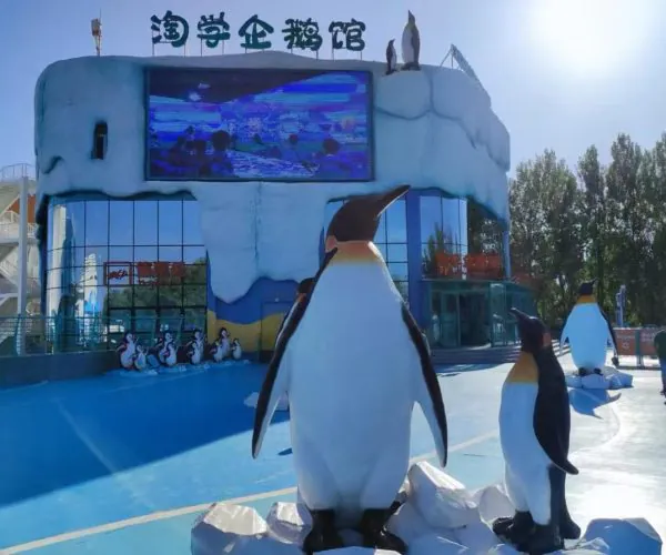 Tall penguin statue at an icy penguin exhibit entrance, with a blue snow-themed building and smaller penguin figures nearby.