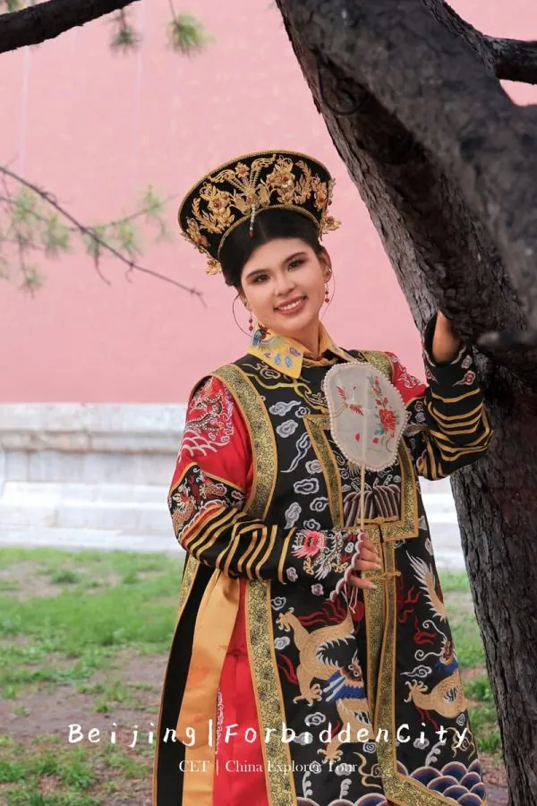 hanfu photoshoot, the forbidden city, beijing, qing dynasty, female, solo, chinatrip, Woman in ornate traditional Chinese imperial robe and headdress, leaning on a tree with a pink wall background.