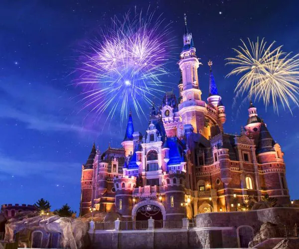 Shanghai-Disneyland-Castle, china, chinaexplorertour, Castle illuminated at night with colorful fireworks bursting overhead, set on a rocky base and blue sky.