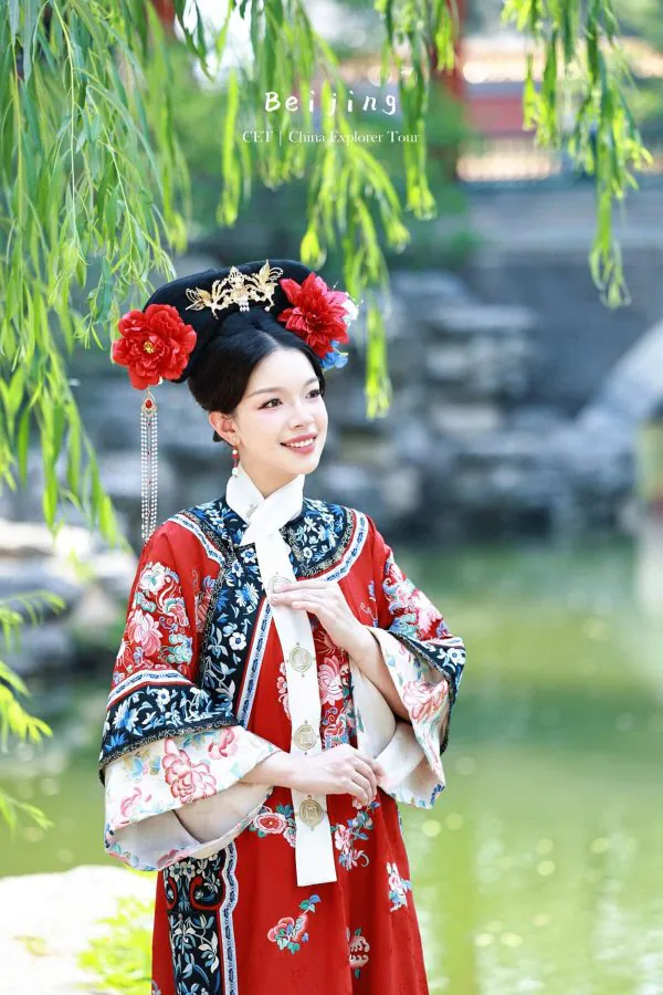 hanfu photoshoot, the forbidden city, beijing, qing dynasty, female, solo, chinatravel, Woman in a red, blue, and white traditional Chinese outfit with an ornate headdress, standing by a pond under hanging willow branches during a Beijing tour.