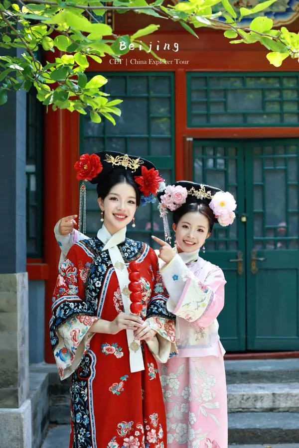 hanfu photoshoot, the forbidden city, beijing, qing dynasty, female, friends, chinatravel, Two smiling women in ornate traditional Chinese dresses with large floral headpieces pose in front of a red doorway with green windows, depicting Beijing attire.