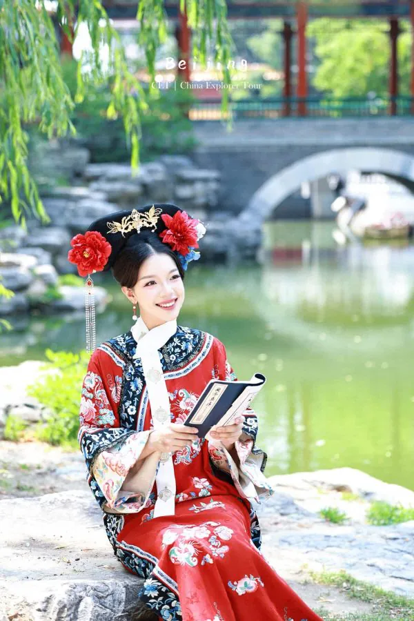 hanfu photoshoot, the forbidden city, beijing, qing dynasty, female, solo, chinatravel, Woman in a red traditional Chinese dress with ornate headpiece reading a book by a pond, bridge in the background