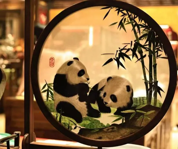 Two pandas sit together among bamboo inside a round framed artwork, warm indoor lighting behind it.