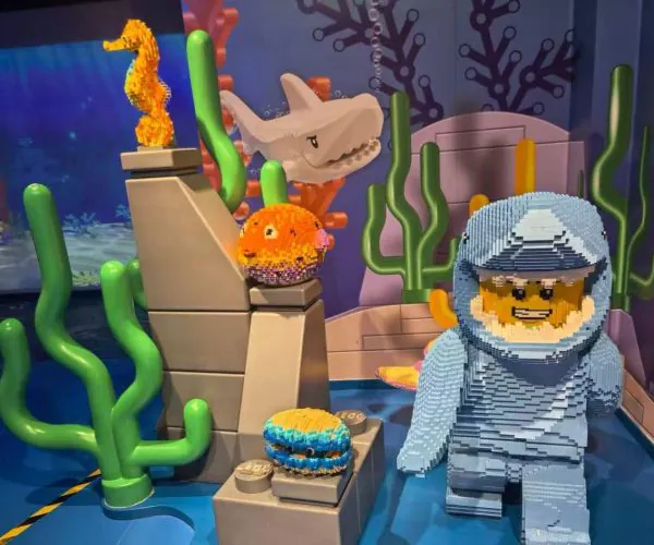 Underwater LEGO scene with a blue-suited scuba diver, a smiling shark, orange fish, and coral in a colorful display.