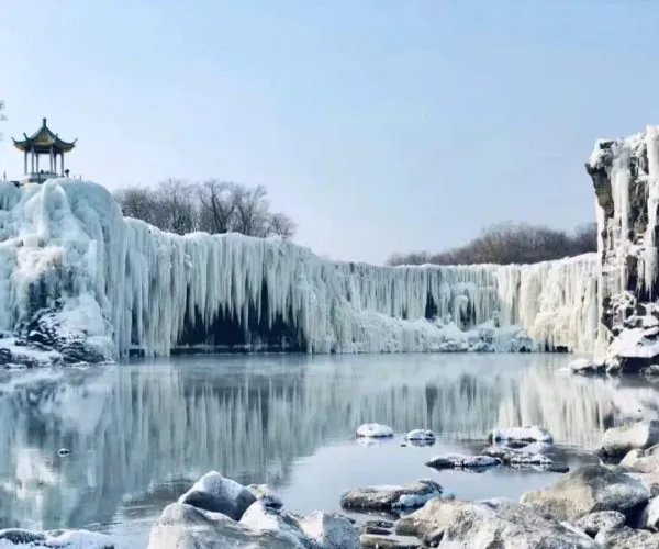 Winter scene with a frozen waterfall and icicles, a small pagoda on the left, and a calm, reflective lake below.