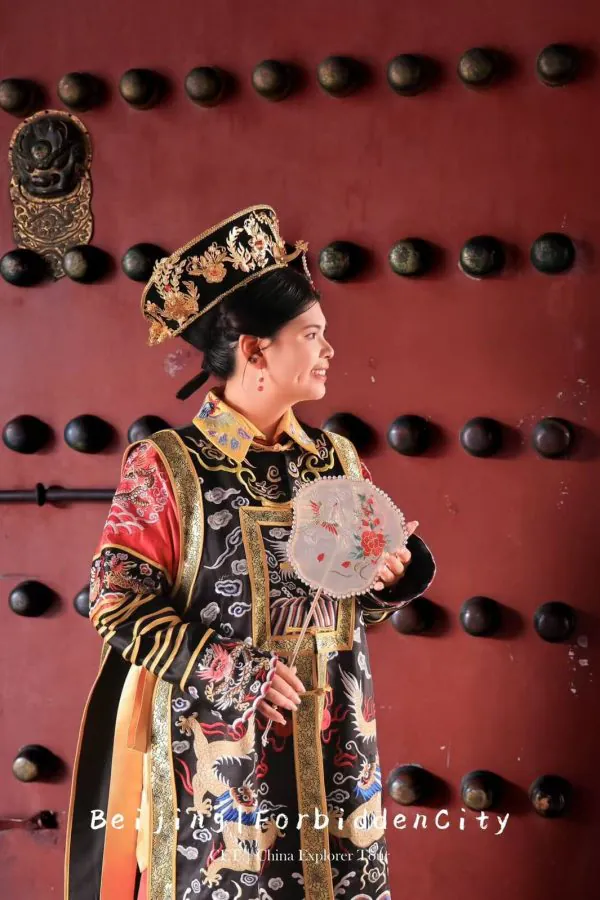 hanfu photoshoot, the forbidden city, beijing, qing dynasty, female, solo, chinatrip, Young woman in an ornate Chinese imperial costume with a jeweled headdress, holding a lace-edged fan against a red studded door.