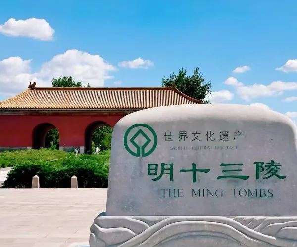 Stone monument with green emblem and Chinese text reading 'World Cultural Heritage' and 'The Ming Tombs' in front of a red gate with tiled roof under a blue sky.