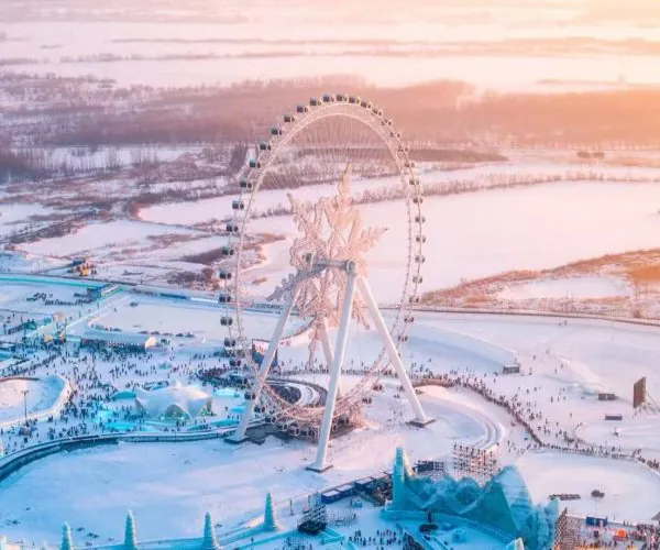 Aerial view of a large, illuminated Ferris wheel in a snow-covered amusement park at sunset, with crowds below.