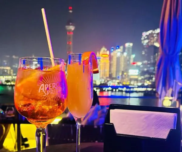 Two cocktails on a rooftop bar at night with a glowing city skyline in the background.