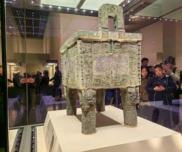 National Museum of China, beijing, chinatravel, Ancient bronze ding with four legs on display in a museum, ornate geometric patterns and a lid, visitors visible in the background behind glass shields.