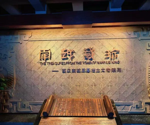 Exhibit wall with intricate geometric carvings and the title 'The Treasures from the Tomb of Manyue King' in English; a wooden display platform sits at the front.
