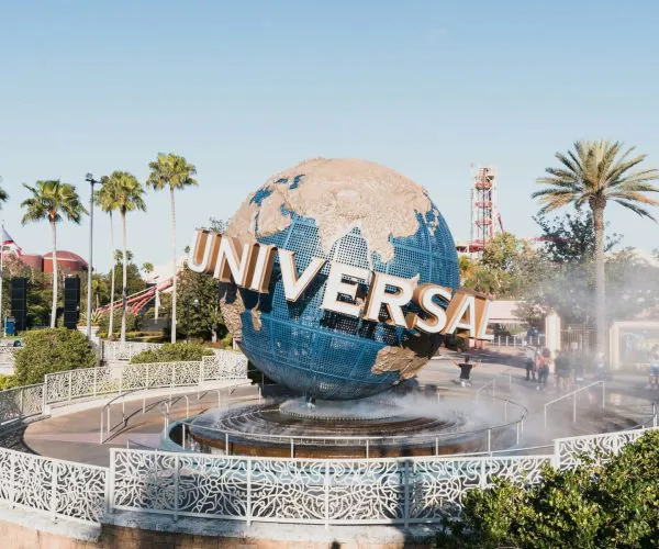 Globe sculpture with blue oceans and gold landmasses, spelling 'UNIVERSAL' in large letters, at Universal Studios theme park entrance with palm trees and mist fountain in foreground.