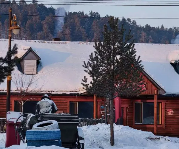 Snowy winter scene at a wooden lodge with a red facade, smoke rising from a chimney, and a person in a heavy coat standing near tires and a bench in the foreground.