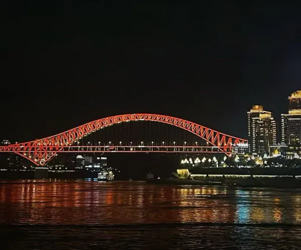 Night view of a red-lit arched bridge spanning a river, with a city skyline illuminated in the background