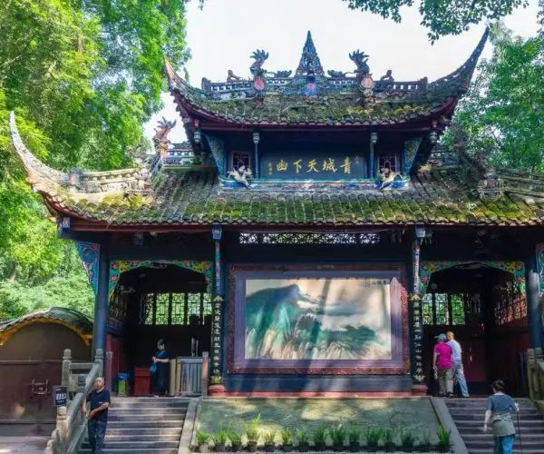 Ornate traditional Chinese pavilion in a garden, with a large mural on the front wall and visitors on the steps.