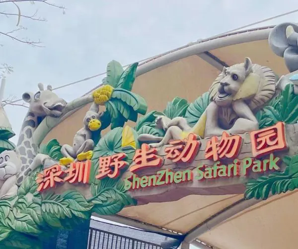 Entrance arch of Shenzhen Safari Park with colorful animal sculptures and tropical foliage (giraffe, monkey, birds) above the sign in Chinese characters and English text.